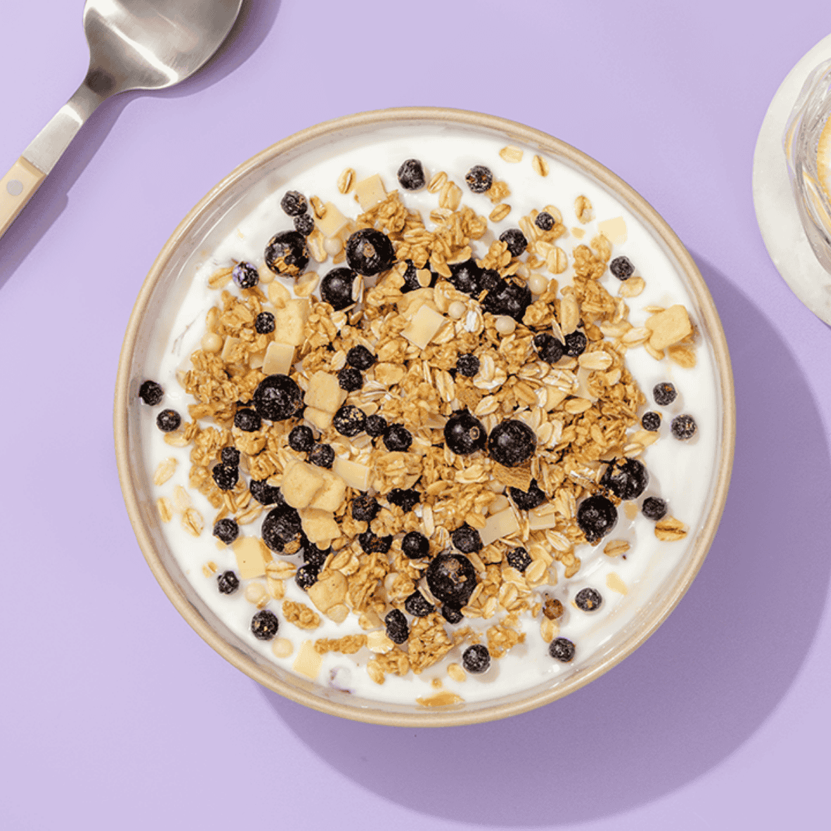 Eine Schüssel mit Müsli und Schokoladen-Crisps auf weißem Joghurt, von oben fotografiert auf einem hellvioletten Hintergrund.