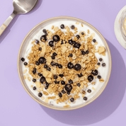 Eine Schüssel mit Müsli und Schokoladen-Crisps auf weißem Joghurt, von oben fotografiert auf einem hellvioletten Hintergrund.