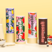 Vier verschiedene mymuesli -Dosen sind dekorativ auf kleinen goldenen Podesten vor einem hellgelben Hintergrund aufgestellt.