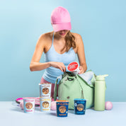 Eine Sportlerin mit pinker Kappe packt einen mymuesli Portionsbecher in ihre grüne Tasche; weitere Müsli-Produkte stehen daneben.