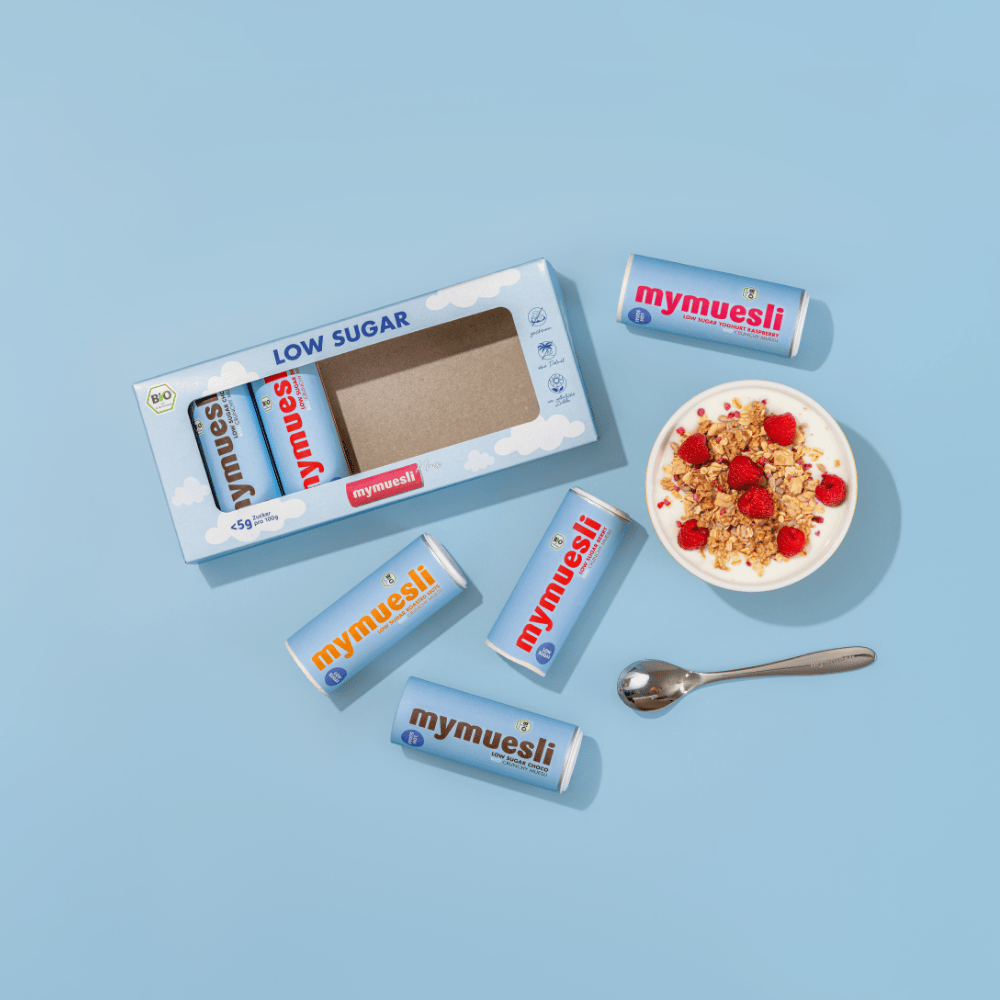 Eine Probiermix-Box, drei Müslischalen und lose verteilte Müsliriegel sowie ein Löffel auf einem hellblauen Hintergrund.