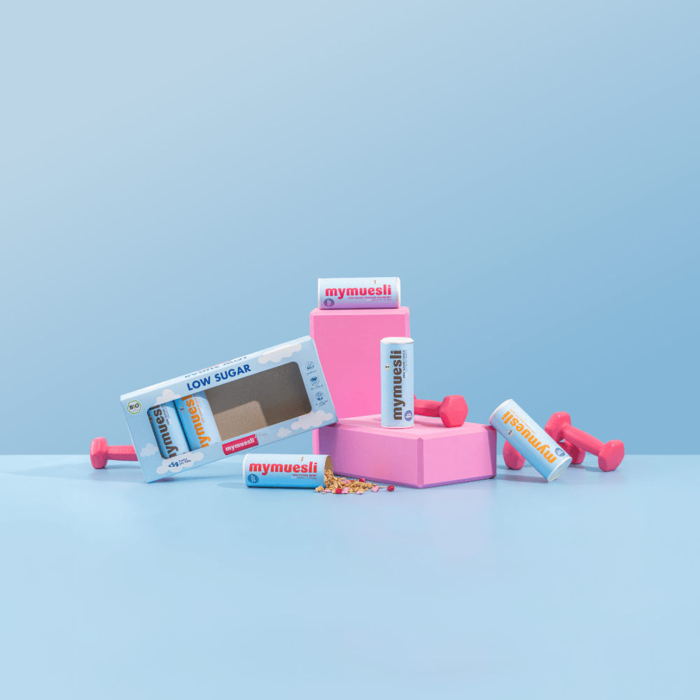 Stillleben mit Müsliriegeln, einer rosa Box und kleinen hantelförmigen Gewichten auf hellblauem Grund – passend zum Thema Fitness.