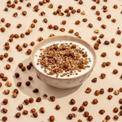 Eine Schüssel mit Joghurt und dunklem Müsli, platziert inmitten vieler kleiner Schokoladen-Knusperstücke auf beigem Untergrund.
