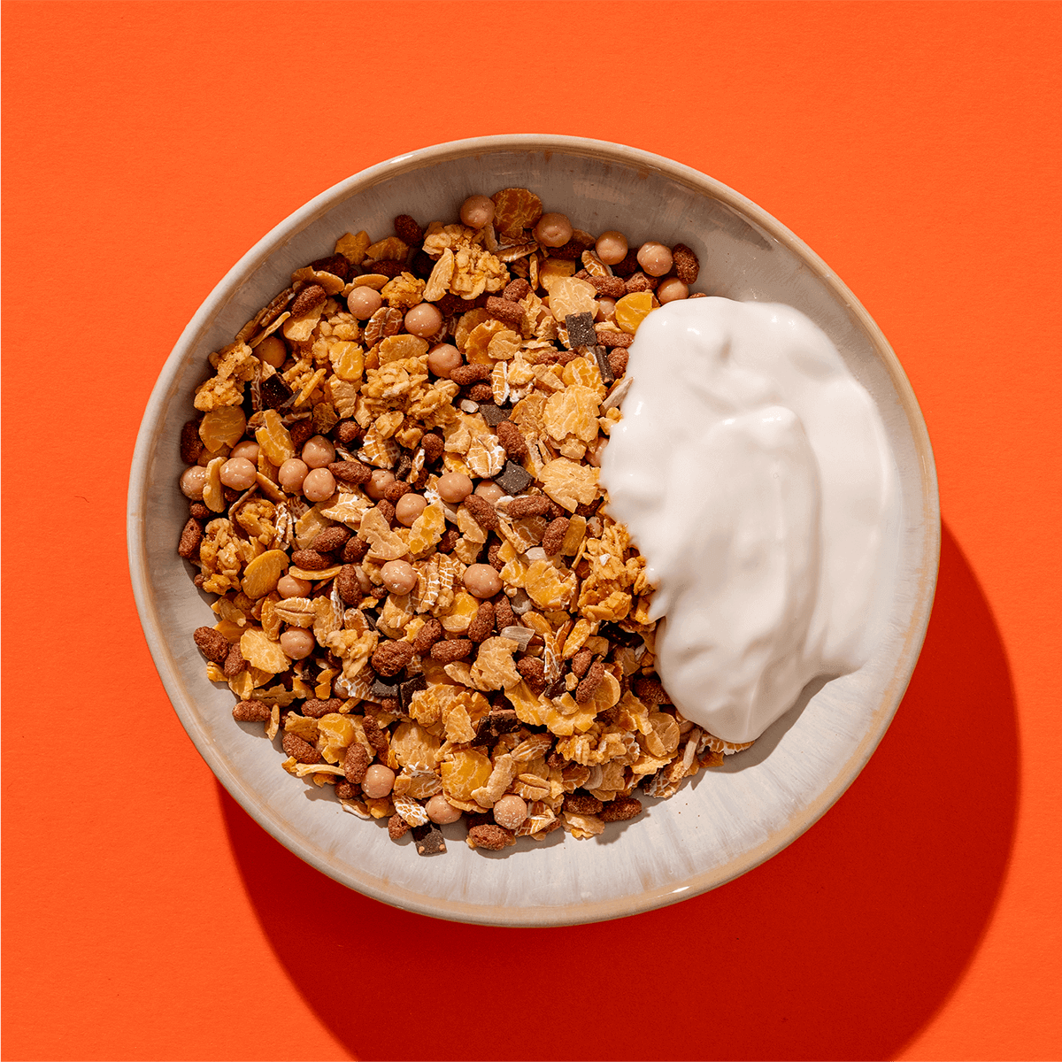 Eine helle Schüssel mit goldbraunem Knusper-Granola und einem Klecks Joghurt vor einem kräftigen orangefarbenen Hintergrund.