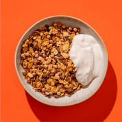 Eine helle Schüssel mit goldbraunem Knusper-Granola und einem Klecks Joghurt vor einem kräftigen orangefarbenen Hintergrund.