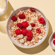 Eine Schale Müsli mit frischen Himbeeren, weißen Schokoladenplättchen und hellen Knusperstücken auf beigem Untergrund.