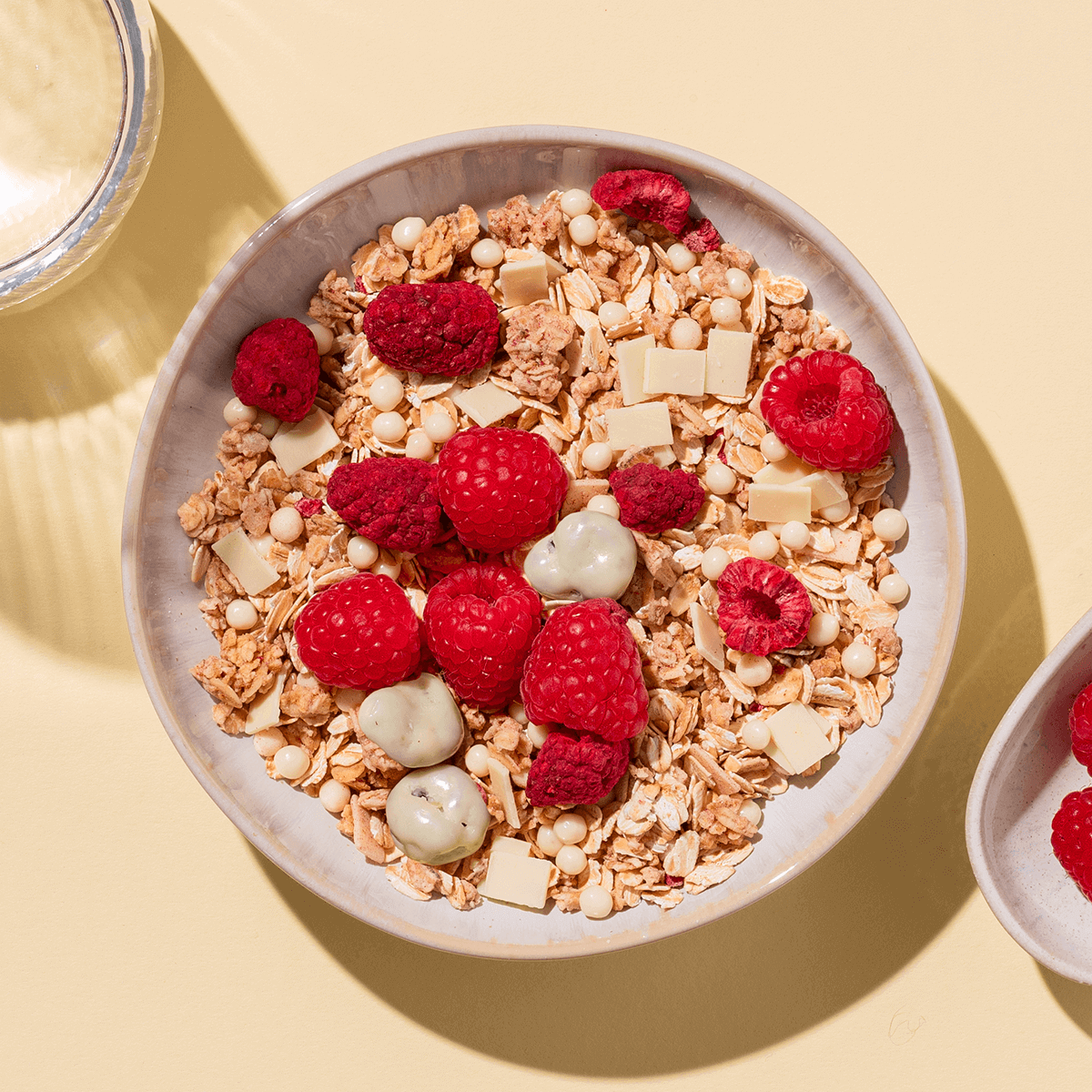 Müsli-Schale mit frischen Himbeeren, weißen Schokoladenstücken und Joghurt-Drops auf hellem, beigem Hintergrund.