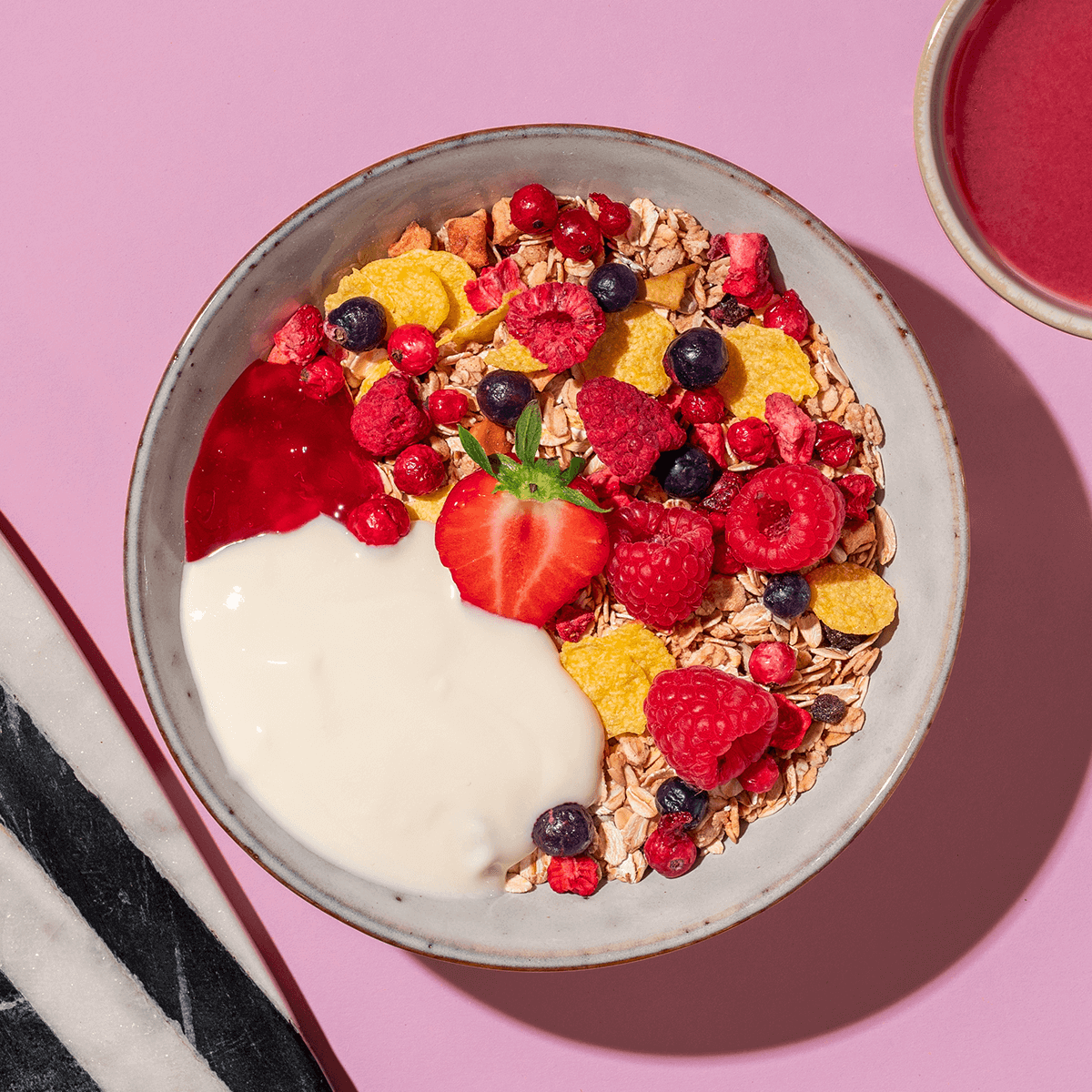 Eine bunte Frühstücksschüssel mit Joghurt, Müsli, Himbeeren und einer halben Erdbeere in der Mitte auf einem rosa-weiß gestreiften Untergrund.