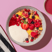 Eine bunte Frühstücksschüssel mit Joghurt, Müsli, Himbeeren und einer halben Erdbeere in der Mitte auf einem rosa-weiß gestreiften Untergrund.