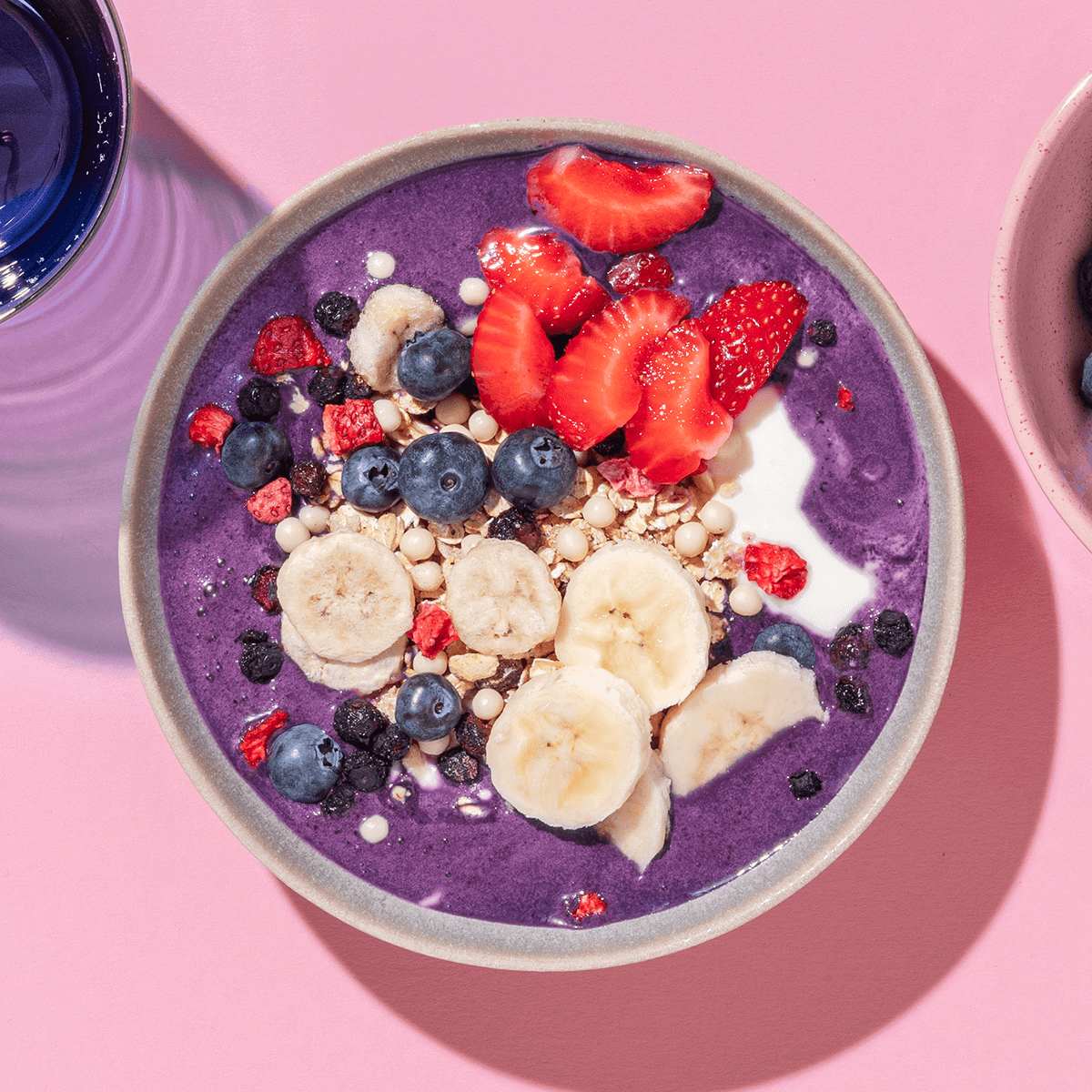 Eine violette Smoothie-Bowl in einer grauen Schüssel, garniert mit Bananenscheiben, Blaubeeren und halbierten Erdbeeren auf einem rosa Hintergrund.