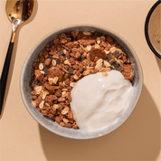 Eine Schüssel mit Schokoladenmüsli und einem Klecks weißem Joghurt, von oben fotografiert auf beigem Untergrund neben einem goldenen Löffel.