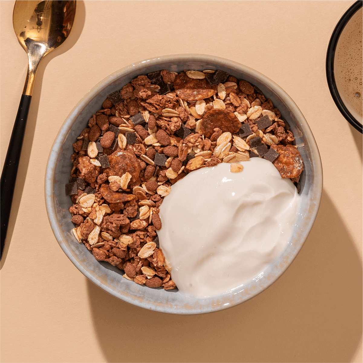 Draufsicht einer Schüssel mit Schokoladenmüsli und einem großen Klecks weißem Joghurt auf sandfarbenem Untergrund.