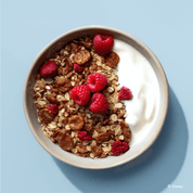 Eine Schüssel mit dunklerem Schoko- oder Knuspermüsli, kombiniert mit Joghurt und frischen Himbeeren auf einem hellblauen Hintergrund.