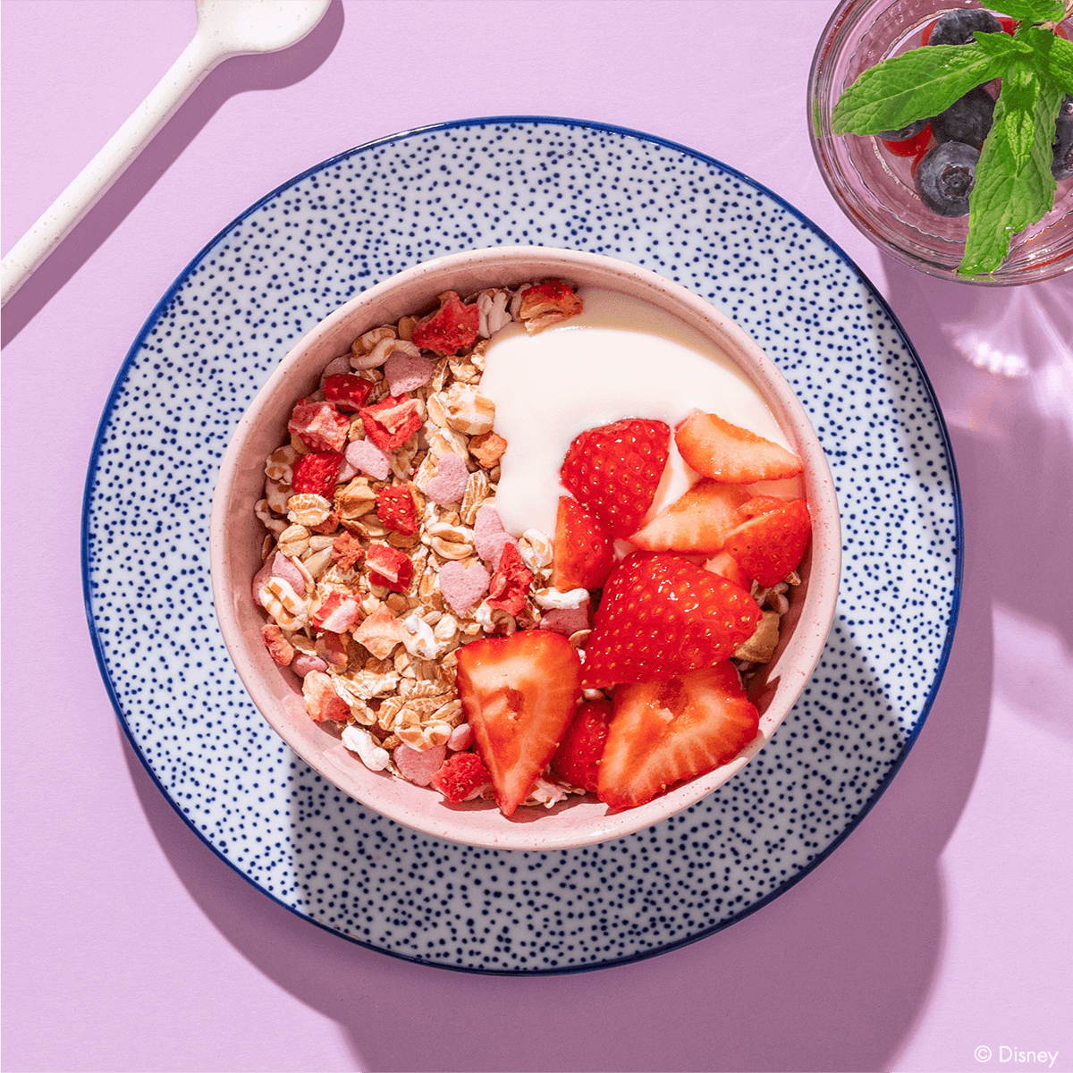 Eine Frühstücksschüssel mit Müsli, Joghurt und Erdbeeren, angerichtet auf einem blau-weiß gemusterten Teller vor einem fliederfarbenen Hintergrund.