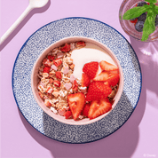 Eine Frühstücksschüssel mit Müsli, Joghurt und Erdbeeren, angerichtet auf einem blau-weiß gemusterten Teller vor einem fliederfarbenen Hintergrund.