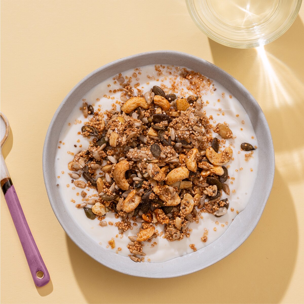 Eine hellblaue Schüssel mit Granola und Milch auf einem sandfarbenen Untergrund, daneben ein Löffel mit lila Griff.