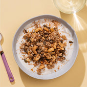 Eine hellblaue Schüssel mit Granola und Milch auf einem sandfarbenen Untergrund, daneben ein Löffel mit lila Griff.