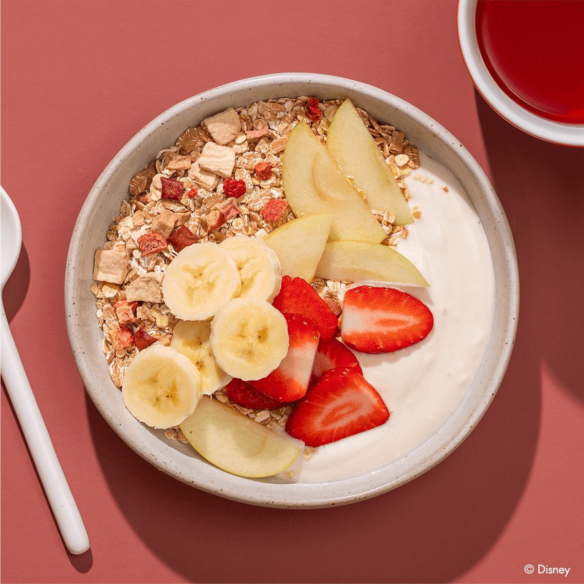 Eine helle Frühstücksschüssel auf rötlichem Untergrund, gefüllt mit Joghurt, Müsli, Bananenscheiben, Erdbeerstücken und Apfelspalten.