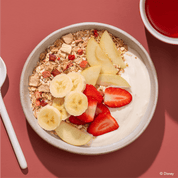 Eine helle Frühstücksschüssel auf rötlichem Untergrund, gefüllt mit Joghurt, Müsli, Bananenscheiben, Erdbeerstücken und Apfelspalten.