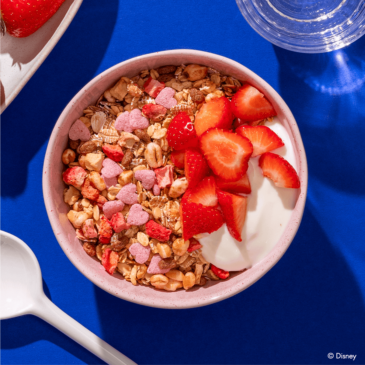 Eine rosafarbene Schüssel mit Müsli, weißem Joghurt und frischen Erdbeerscheiben, garniert mit kleinen rosa Herzchen auf einem leuchtend blauen Hintergrund.