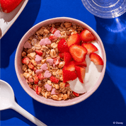 Eine rosafarbene Schüssel mit Müsli, weißem Joghurt und frischen Erdbeerscheiben, garniert mit kleinen rosa Herzchen auf einem leuchtend blauen Hintergrund.