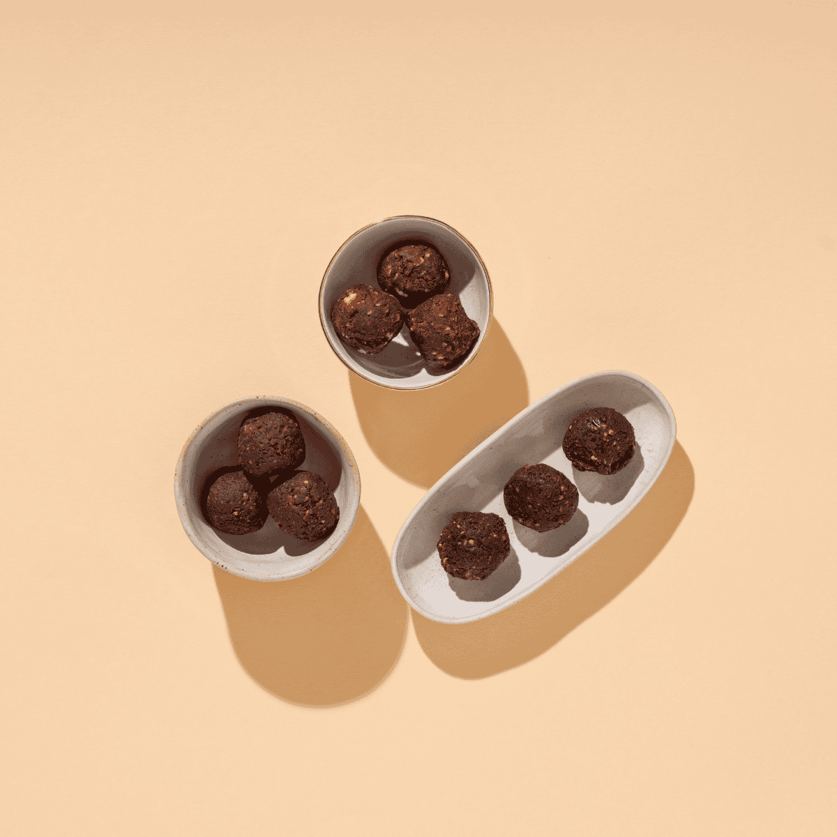 Drei kleine Schälchen auf beigem Grund, die mit dunklen Schokoladenkugeln (Nut Butter Balls) gefüllt sind.