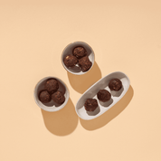 Drei kleine Schälchen auf beigem Grund, die mit dunklen Schokoladenkugeln (Nut Butter Balls) gefüllt sind.