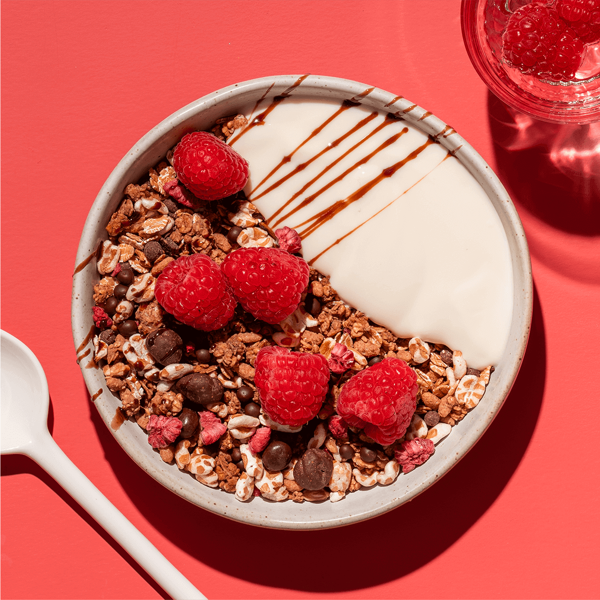 Eine Schüssel mit Schokoladenmüsli, Joghurt und frischen Himbeeren auf rotem Grund, verziert mit feinen Schokoladenlinien.
