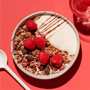 Eine Schüssel mit Schokoladenmüsli, Joghurt und frischen Himbeeren auf rotem Grund, verziert mit feinen Schokoladenlinien.