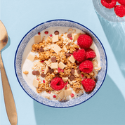 Eine blau-weiß gemusterte Schüssel mit Müsli und Himbeeren, von oben fotografiert auf hellblauem Untergrund neben einem goldenen Löffel.