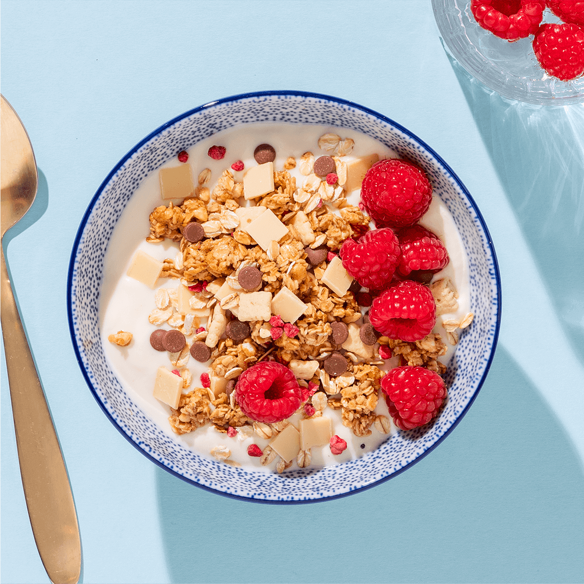 Eine blau-weiß gemusterte Schüssel mit Müsli und Himbeeren, von oben fotografiert auf hellblauem Untergrund neben einem goldenen Löffel.