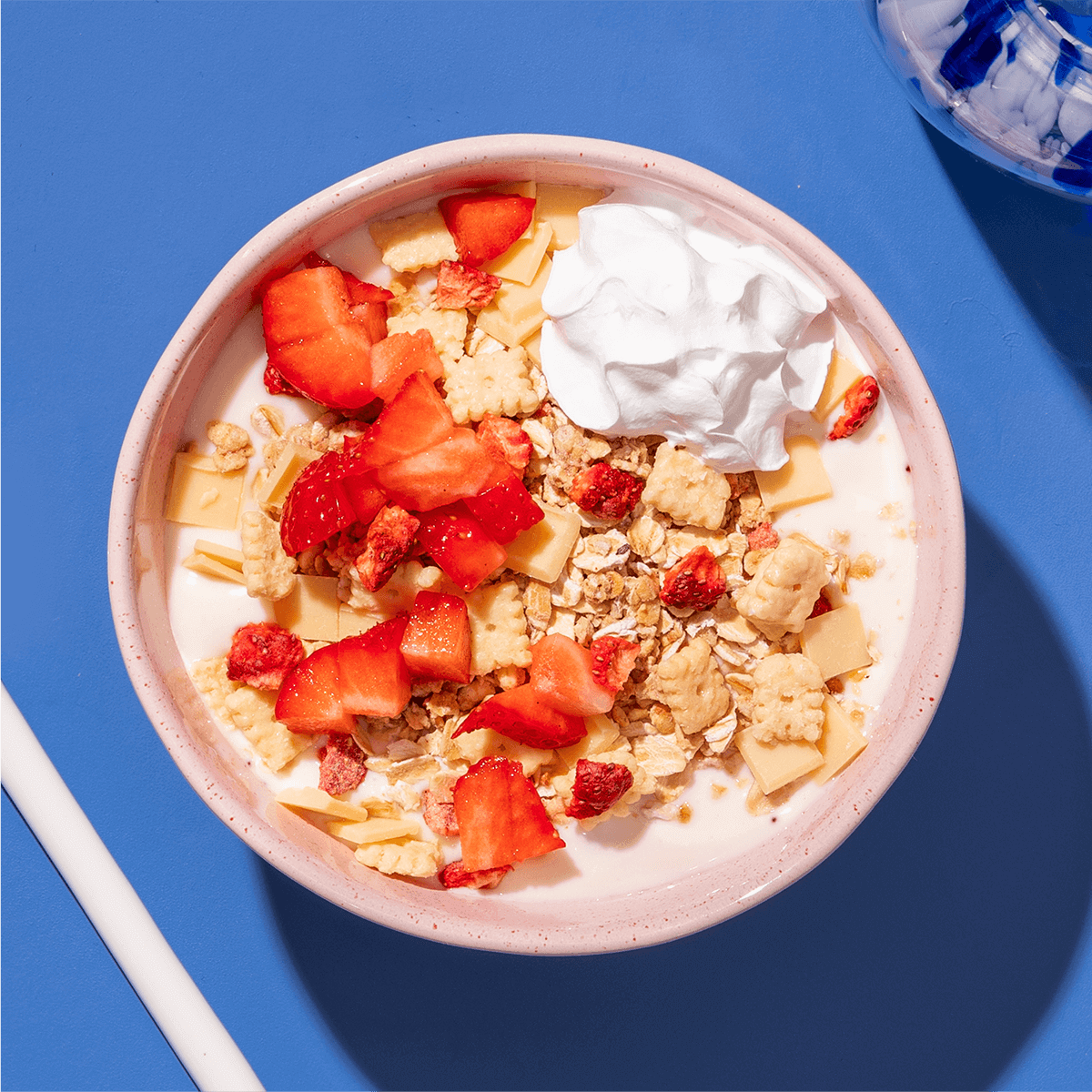 Eine rosa Schüssel mit hellem Müsli, Erdbeerstücken und einem Klecks Sahne oder Joghurt auf einem blauen Untergrund.