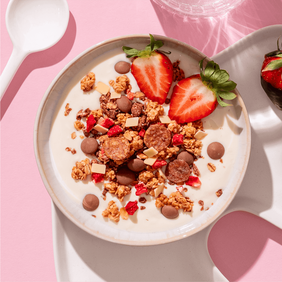 Eine Schüssel mit Müsli, Joghurt und halbierten Erdbeeren auf einem rosa Hintergrund, daneben liegt ein weißer Löffel.