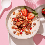 Eine Schüssel mit Müsli, Joghurt und halbierten Erdbeeren auf einem rosa Hintergrund, daneben liegt ein weißer Löffel.