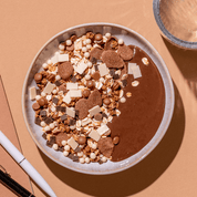 Draufsicht einer Müsli-Schale mit Schokoladen-Granola, dunklen Schokoplättchen und Kokoschips auf beigem Hintergrund.