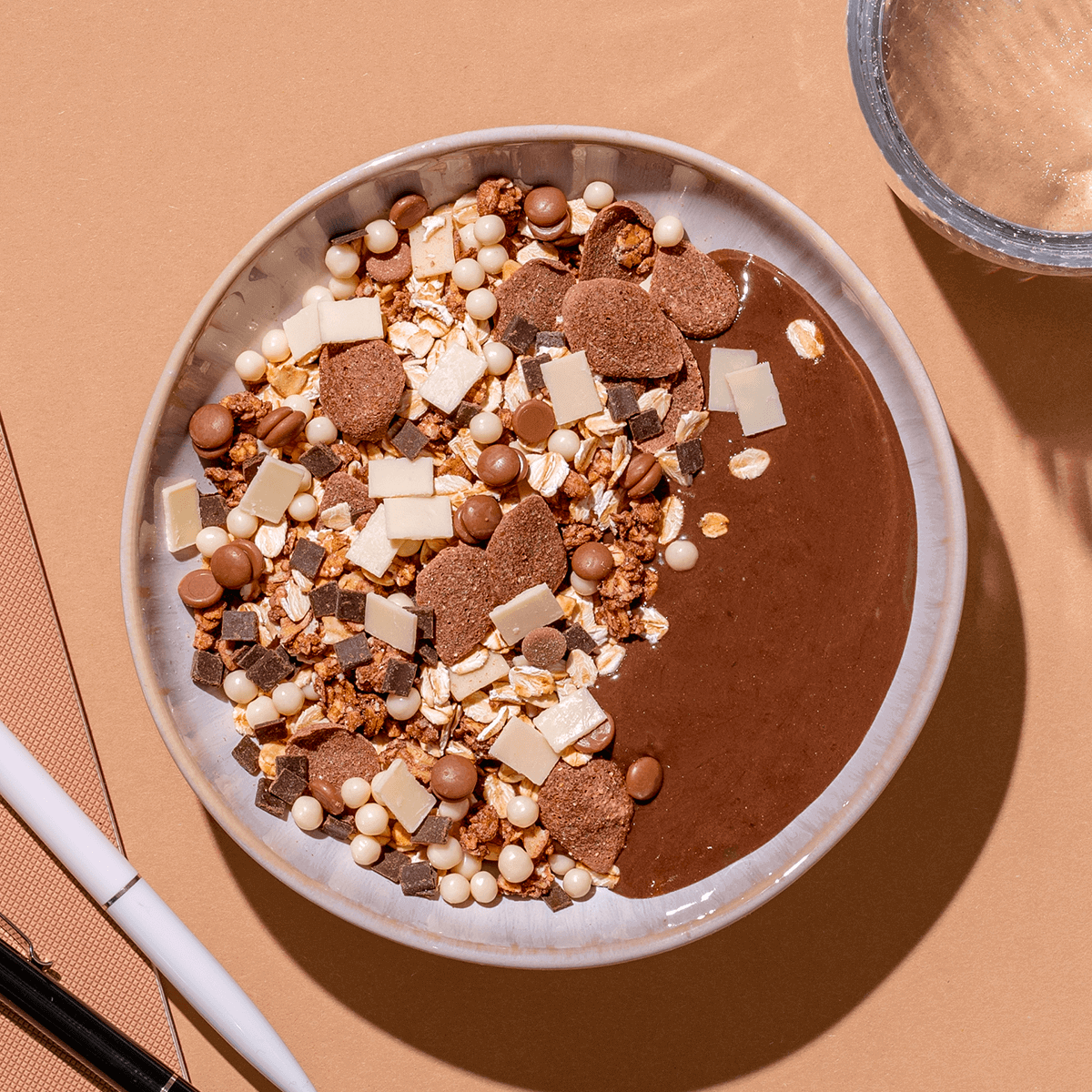 Draufsicht einer Müsli-Schale mit Schokoladen-Granola, dunklen Schokoplättchen und Kokoschips auf beigem Hintergrund.