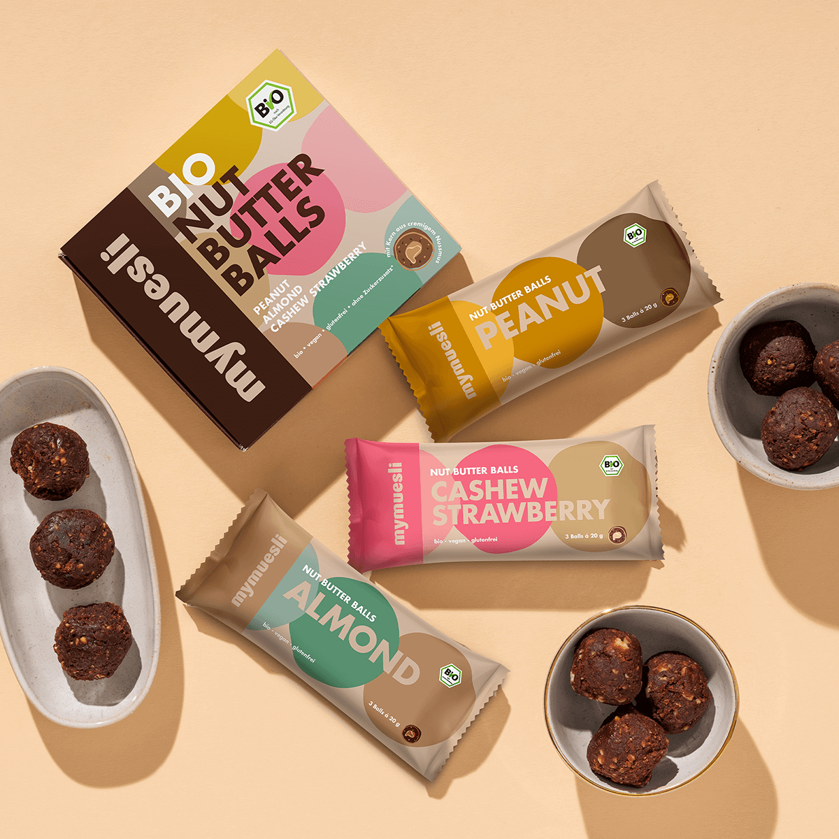 Ein Stillleben mit einer Packung "Nut Butter Balls", verschiedenen Müsliriegeln (Peanut, Strawberry, Almond) und kleinen Schalen mit Schokoladenkugeln auf beigem Grund.