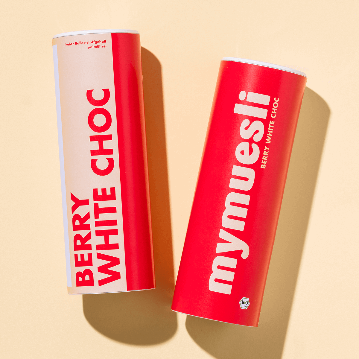 Zwei mymuesli -Dosen der Sorte 'Berry White Choc' in Weiß und Rot auf einem sandfarbenen Untergrund.