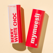 Zwei mymuesli -Dosen der Sorte 'Berry White Choc' in Weiß und Rot auf einem sandfarbenen Untergrund.