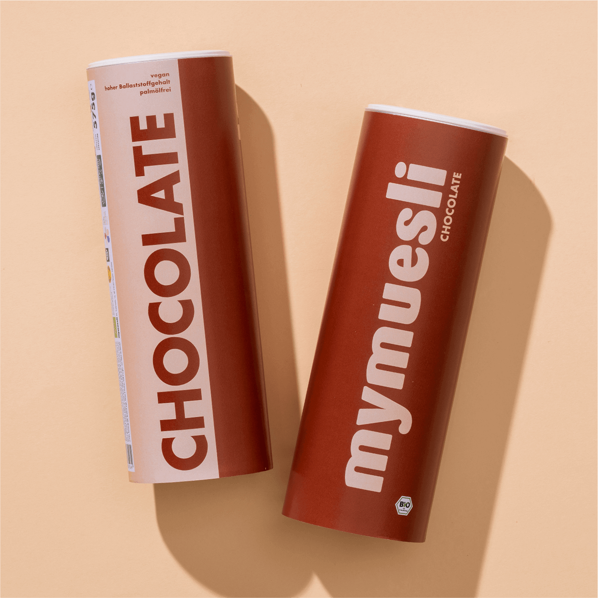 Zwei mymuesli -Dosen der Sorte Chocolate in Braun und Weiß, flach liegend auf beigem Hintergrund.