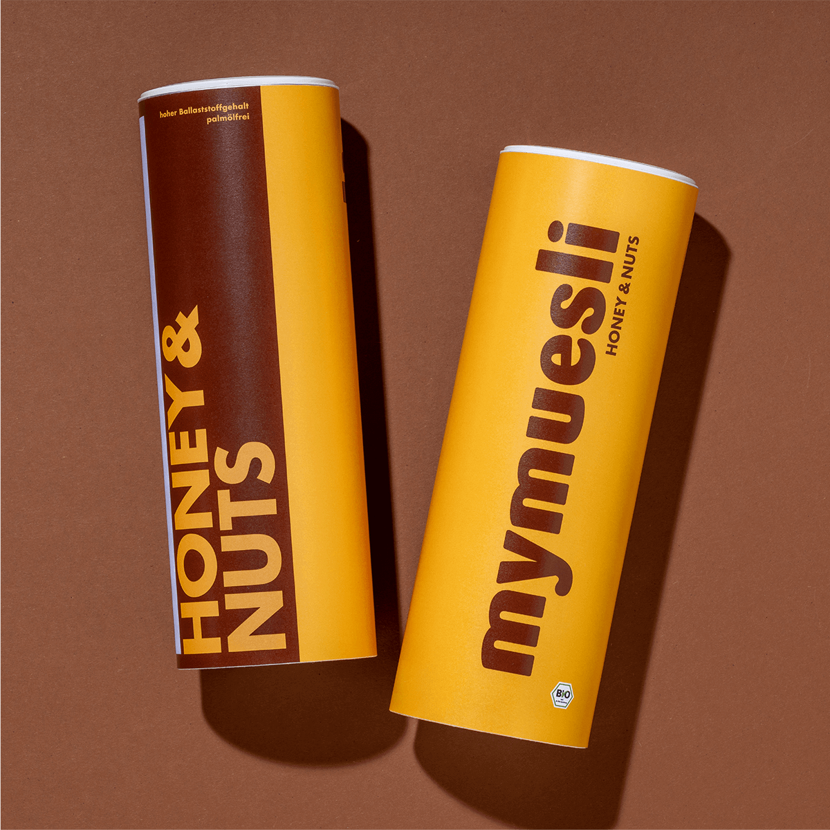 Zwei mymuesli -Dosen der Sorte 'Honey & Nuts' in Braun und Gelb, flach liegend auf braunem Hintergrund.