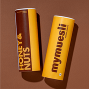 Zwei mymuesli -Dosen der Sorte 'Honey & Nuts' in Braun und Gelb, flach liegend auf braunem Hintergrund.