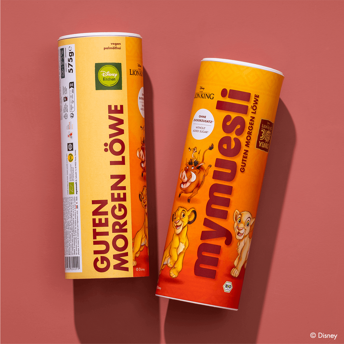 Zwei gelbe mymuesli -Dosen der Edition "Guten Morgen Löwe" mit Disney-Motiven, nebeneinander auf einem rötlich-braunen Hintergrund.