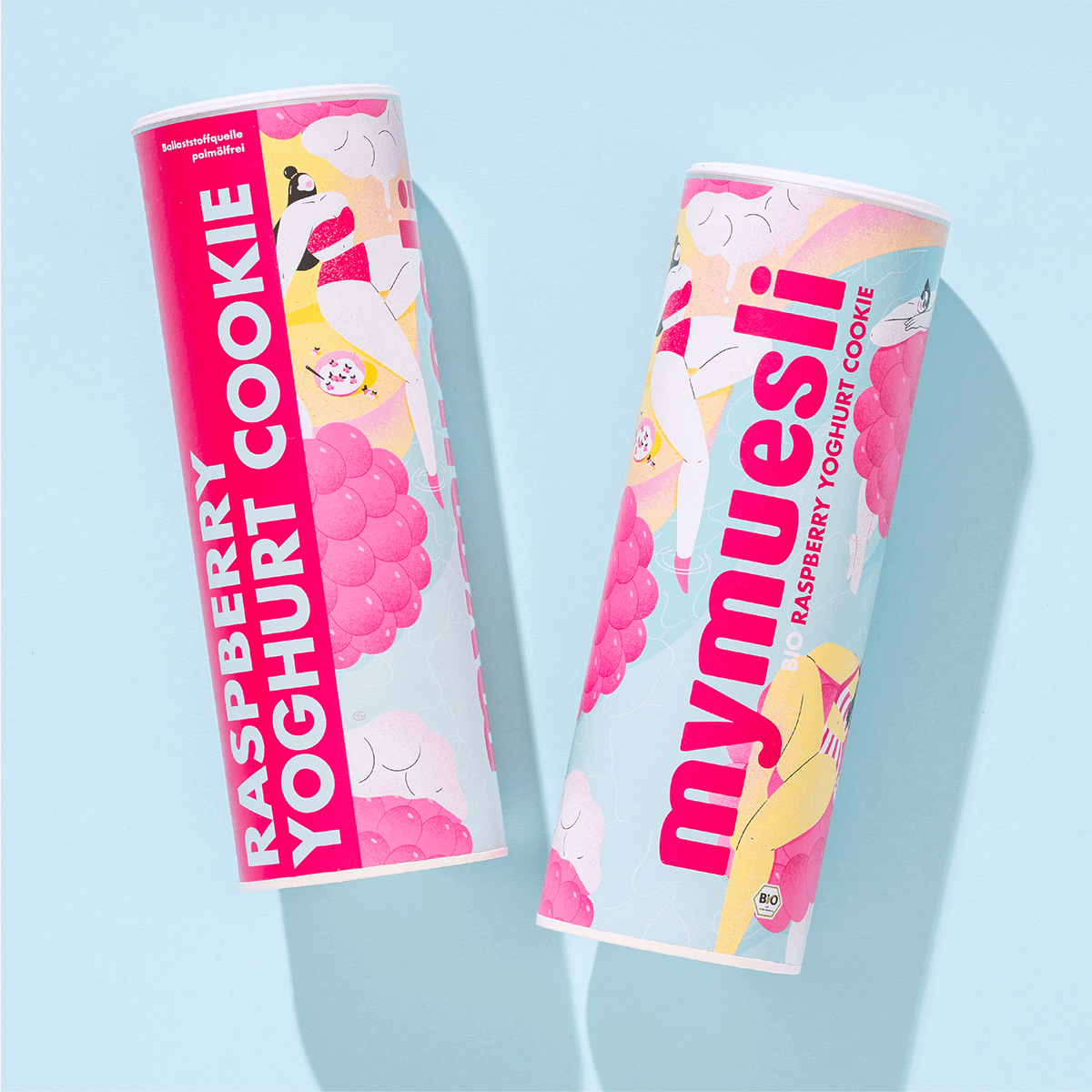 Zwei mymuesli -Dosen der Sorte "Raspberry Yoghurt Cookie" mit pastellfarbenem Design, nebeneinander liegend auf einem hellblauen Hintergrund.