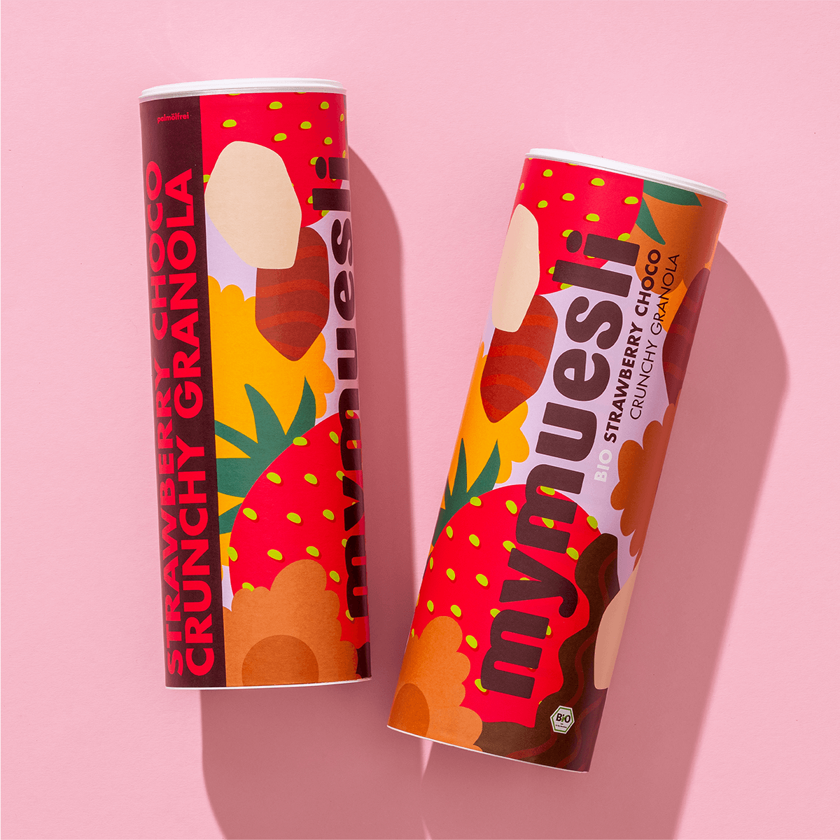 Zwei mymuesli -Dosen mit Erdbeer-Design liegen nebeneinander auf einem hellrosa Hintergrund.