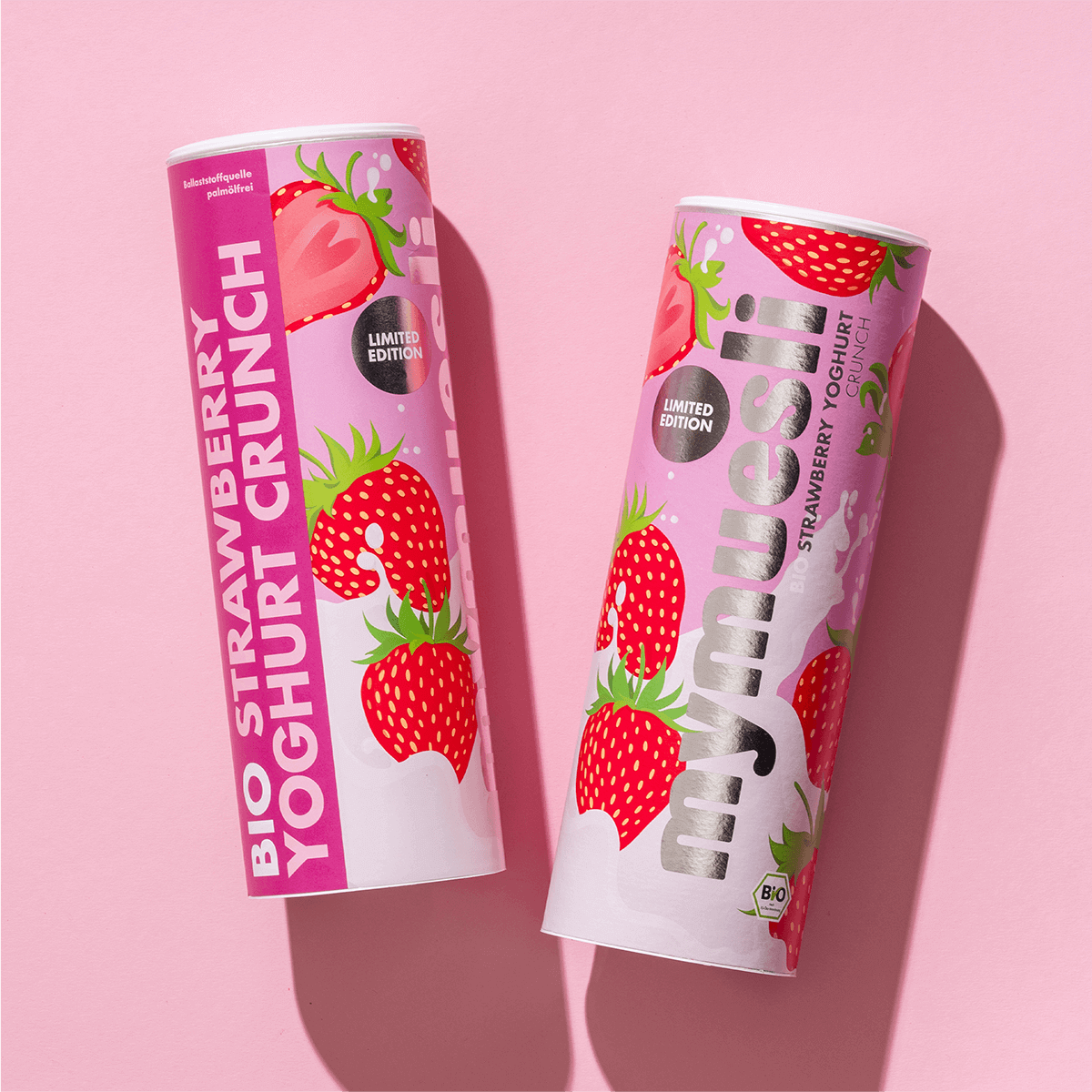 Zwei mymuesli -Dosen der Sorte "Strawberry Yoghurt Crunch" mit Erdbeer-Motiven, nebeneinander liegend auf einem hellrosa Hintergrund.