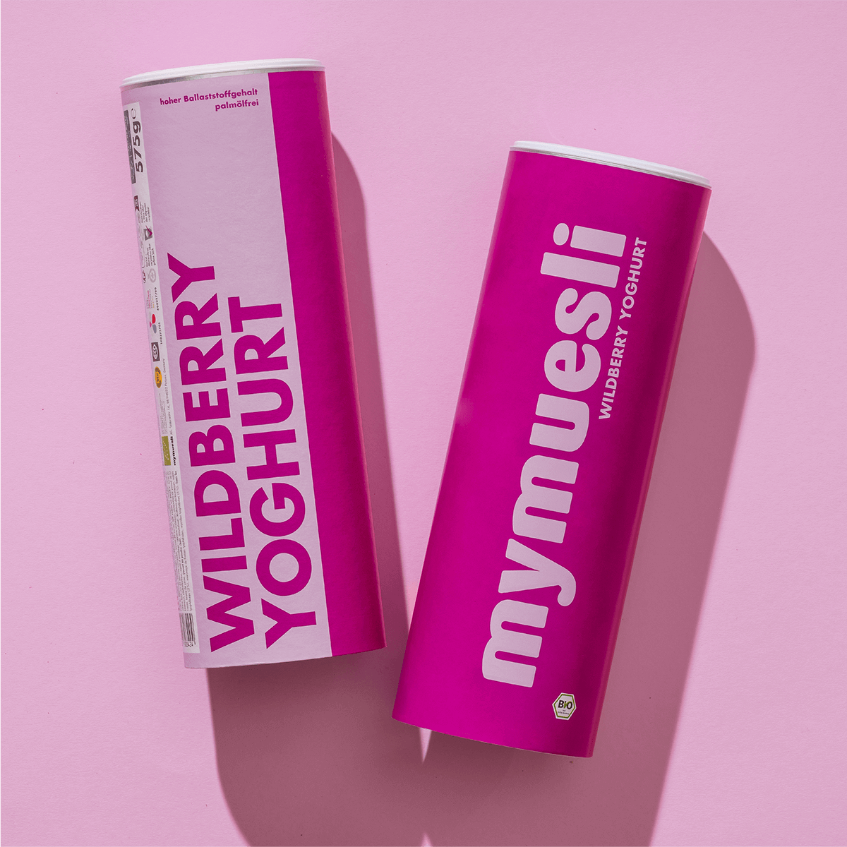 Zwei mymuesli -Dosen der Sorte "Wildberry Yoghurt" – eine hellrosa und eine dunkelpink – liegen nebeneinander auf einem fliederfarbenen Hintergrund.