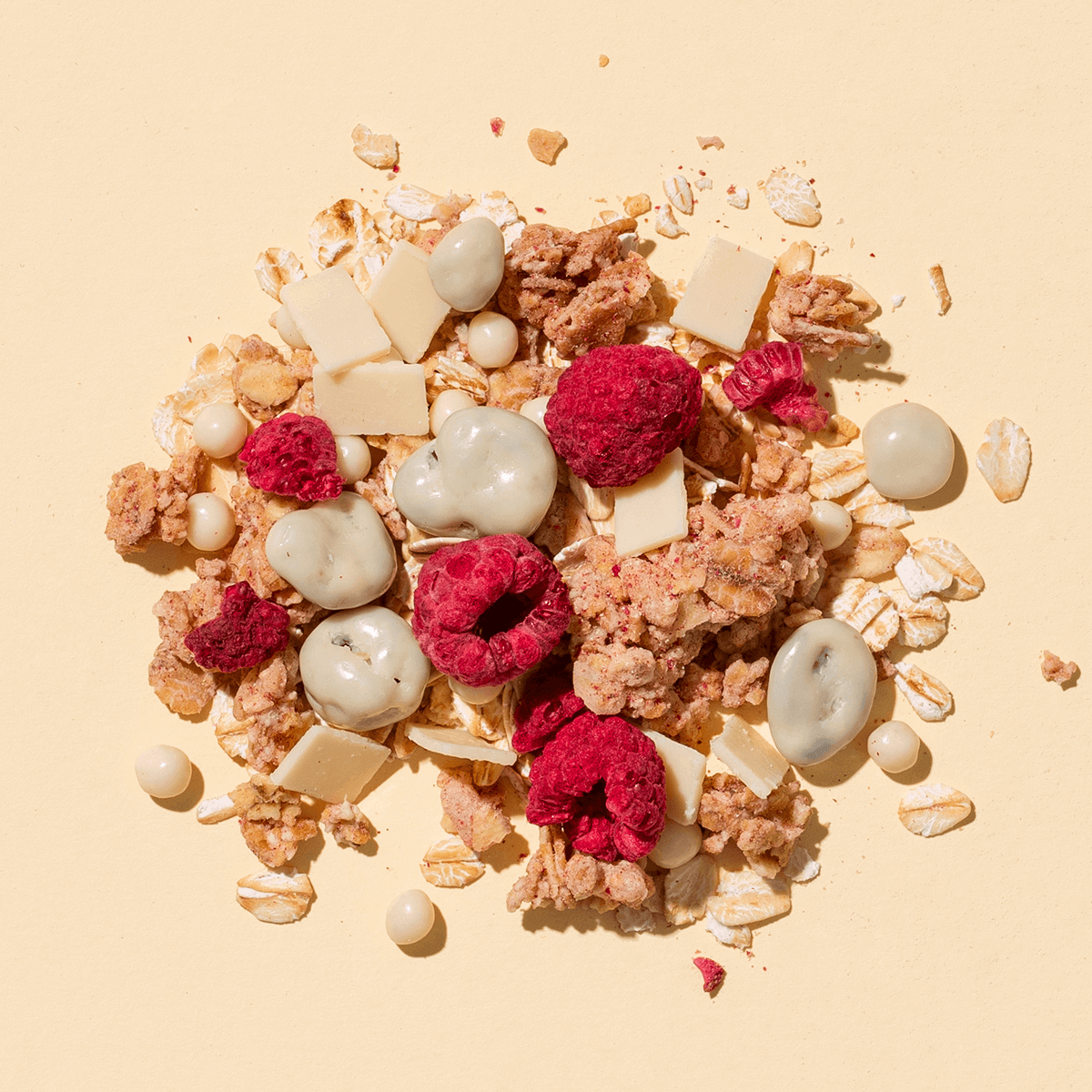 Eine Nahaufnahme der Müsli-Mischung auf beigem Grund mit gefriergetrockneten Himbeeren, weißen Schokostücken und Joghurt-Flakes.
