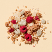Eine Nahaufnahme der Müsli-Mischung auf beigem Grund mit gefriergetrockneten Himbeeren, weißen Schokostücken und Joghurt-Flakes.