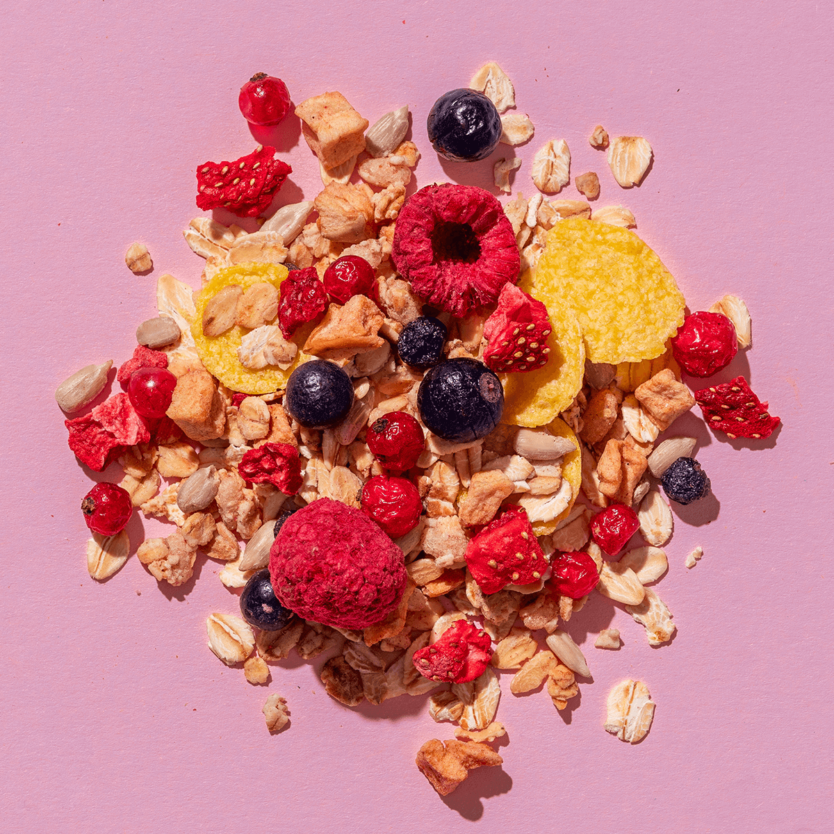 Eine Müsli-Mischung auf rosa Hintergrund mit gefriergetrockneten Himbeeren, Blaubeeren, gelben Fruchtstücken und Haferflocken.